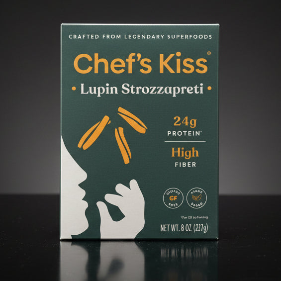 Box of Chef's Kiss Lupin Strozzapreti pasta on a wooden surface with a dark blue fabric background.