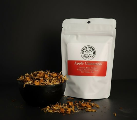White packaging of Apple Cinnamon tea with a black bowl of dried herbs on a dark background