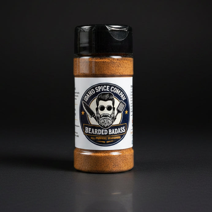 A bottle of Idaho Spice Company Bearded Badass all-purpose seasoning standing on a light wood tabletop with a dark blue curtain in the background.