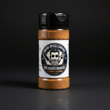 A bottle of Idaho Spice Company Bearded Badass all-purpose seasoning standing on a light wood tabletop with a dark blue curtain in the background.