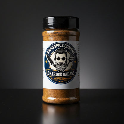 A clear plastic spice shaker bottle filled with brown seasoning, featuring a white label with a cartoon bearded man and the text 'IDAHO SPICE COMPANY' and 'BEARDED BADASS'. The bottle sits on a light wood tabletop with a dark curtain background.