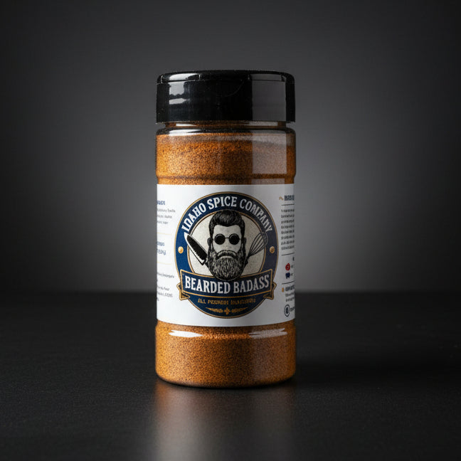 A clear plastic shaker bottle of Idaho Spice Company Bearded Badass seasoning, filled with a brown spice blend, sitting on a light wood tabletop with a dark curtain background.