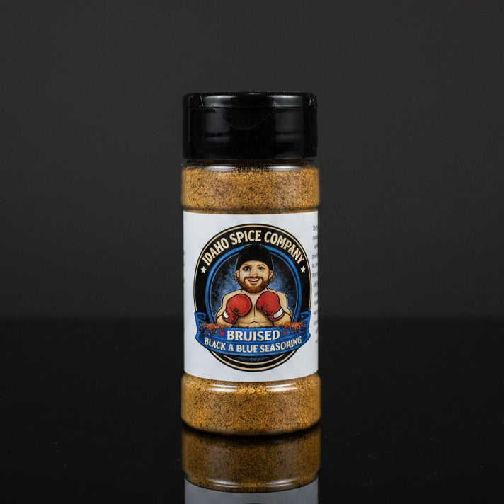 A bottle of Idaho Spice Company Bruised Black & Blue Seasoning, featuring a white label with a cartoon boxer and a black cap, set against a dark reflective background.