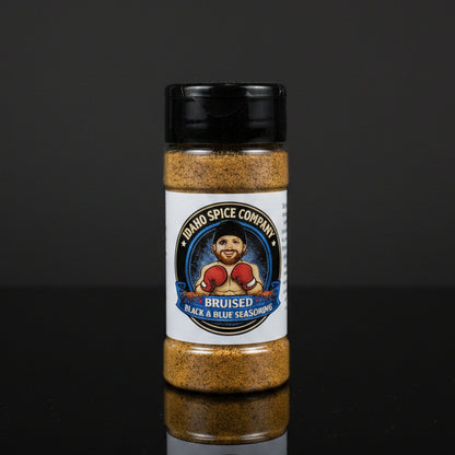 A bottle of Idaho Spice Company Bruised Black & Blue Seasoning, featuring a white label with a cartoon boxer and a black cap, set against a dark reflective background.