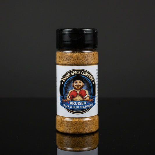 A bottle of Idaho Spice Company Bruised Black & Blue Seasoning, featuring a white label with a cartoon boxer and a black cap, set against a dark reflective background.